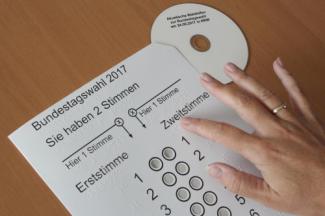 Masada üzerinde Almanca yazılar ve aynı zamanda Braille bulunan beyaz bir oy şablonu ve şablonun sağ üst köşesinde üzerinde yazılar olan bir cd var. İşaret parmağında altın bir yüzük olan bir el Braille noktaları okuyor. Şablonun ortasından aşağıya doğru her birinde yan yana iki yuvarlak boşluğun olduğu bir sütun iniyor.