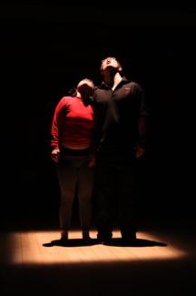 Karanlık bir ortamda birbirilerine değer şekilde yan yana duran ve kafalarını kaldırmış yukarı bakan bir kadın ve bir erkek. Üzerlerine spot ışığı yansıtılmış.