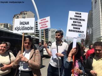 Görme engellilerin işlemlerinde iki tanık istenmesine itiraz için Türkiye Noterler Birliği önünde bir araya gelen görme engellilerin eyleminden bir fotoğraf. Fotoğrafta ellerinde pankart tutan görme engelli kadın ve erkekler yer alıyor. Pankartlarda imzam kişiliğimdir, noter kanundan üstün değildir, yasa tanık istenemezken noter körlere tanık dayatamaz ifadeleri yer alıyor. Arka planda binalar ve araçlar görünüyor. Sol üstte fotoğrafın kaynağı medyascope.tv olarak belirtilmiş.