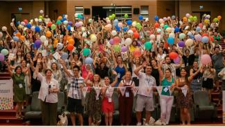 Öğretmen Ağı Değişim Elçileri 2023 Yaz Programından bir fotoğraf. Büyük bir konferans salonu. Koltuklarında ayağa kalkmış, rengârenk balonlarla gülümseyerek ve objektife el sallayarak poz veren kadın erkek onlarca kişi.