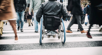 Yaya geçidinde sadece boyundan aşağılarını gördüğümüz ve farklı yönlere doğru hızlıca yürüyen insan kalabalığının tam ortasında sırtı dönük erkek bir tekerlekli sandalye kullanıcısı karşıya geçiyor.