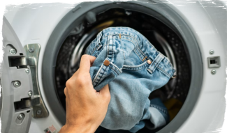 Görselde, kapağı tamamen açık  olan  çamaşır makinasının bir bölümü görüntüleniyor.Bir erkek eli, kot pantolonu çamaşır makinasının içine koyuyor.