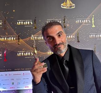 Siyah takım elbise giymiş yönetmen Cüneyt Karakuş Uluslararası Antares Film Festivali panosu önünde aldığı Seyirci Özel Ödülünü omuz hizasına kaldırmış objektife bakıyor. Kır saçları, sakalı ve bıyığı var.