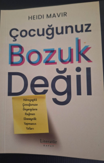 Fotoğrafta bir kitap kapağı görülüyor. Kapağın üst kısmında, "HEIDI MAVIR" yazar ismi siyah renkte yazılmış. Hemen altında, "Çocuğunuz" kelimesi mavi renkte ve ortalanmış bir şekilde yer alıyor. Onun altında ise "Bozuk" kelimesi kırmızı ve mavi renklerin üst üste binmesiyle oluşturulmuş, hafifçe üç boyutlu bir etki yaratıyor. En altta ise "Değil" kelimesi kalın ve siyah harflerle yazılmış. Kitabın tam ortasında, sarı bir post-it yapışkan not var. Notun üzerinde, siyah renkle yazılmış şu metin bulunuyor: