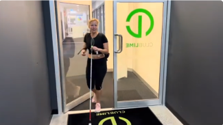 Spor salonu girişinde bir kadın. Kadın, sol elinde beyaz baston tutuyor ve bastonun ucu yerde. Siyah kısa kollu bir tişört ve siyah tayt giymiş, ayaklarında açık pembe spor ayakkabılar var. Saçları açık sarı ve başının üst kısmından geriye doğru toplanmış. Arka planda cam kapılar var; kapıların üzerinde yeşil renkte “CLUB LIME” yazısı ve yuvarlak bir logo dikkat çekiyor. Kadın, kapıdan içeri doğru adım atarken gülümsüyor.