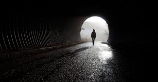 Karanlık tünelden tek başına yürüyerek çıkan bir kadın silueti. Tünelin sonundaki açıklık, parlak bir beyaz ışıkla aydınlanmış durumda. 
