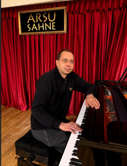 Görselde, bir sahnede piyano başında oturan orta yaşlı bir adam yer alıyor.  İnce çerçeveli gözlük takan, kısa saçlı, hafif gülümseyen bir adam; üzerinde siyah, fermuarlı bir hırka ve gri pantolon var. Adam siyah bir piyanonun taburesinde oturuyor. Sağ eli piyanonun tuşları üzerinde, sol eli ise piyanonun gövdesine dayanmış durumda. Sahnenin arkasında boydan boya uzanan, dikey kıvrımlı, canlı kırmızı renkte bir perde bulunuyor. Perdenin üzerinde, altın sarısı çerçeveli siyah bir tabela asılı.