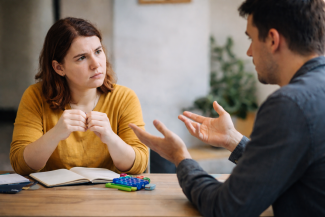 Bir masa başında karşılıklı oturan iki yetişkin görülüyor. Soldaki kişi dikkatle dinlerken ellerini bir arada tutuyor; sağdaki kişi konuşur gibi elleriyle açıklayıcı bir hareket yapıyor. Masada açık bir defter ve renkli küçük nesneler bulunuyor.
